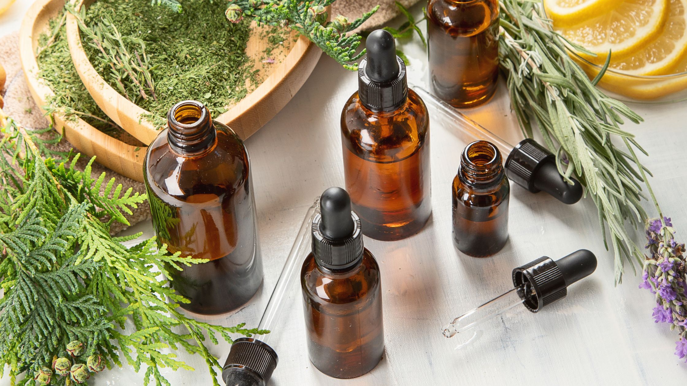 Assortment of amber essential oil bottles with dropper caps, surrounded by fresh herbs, dried leaves, and lemon slices on a white surface.