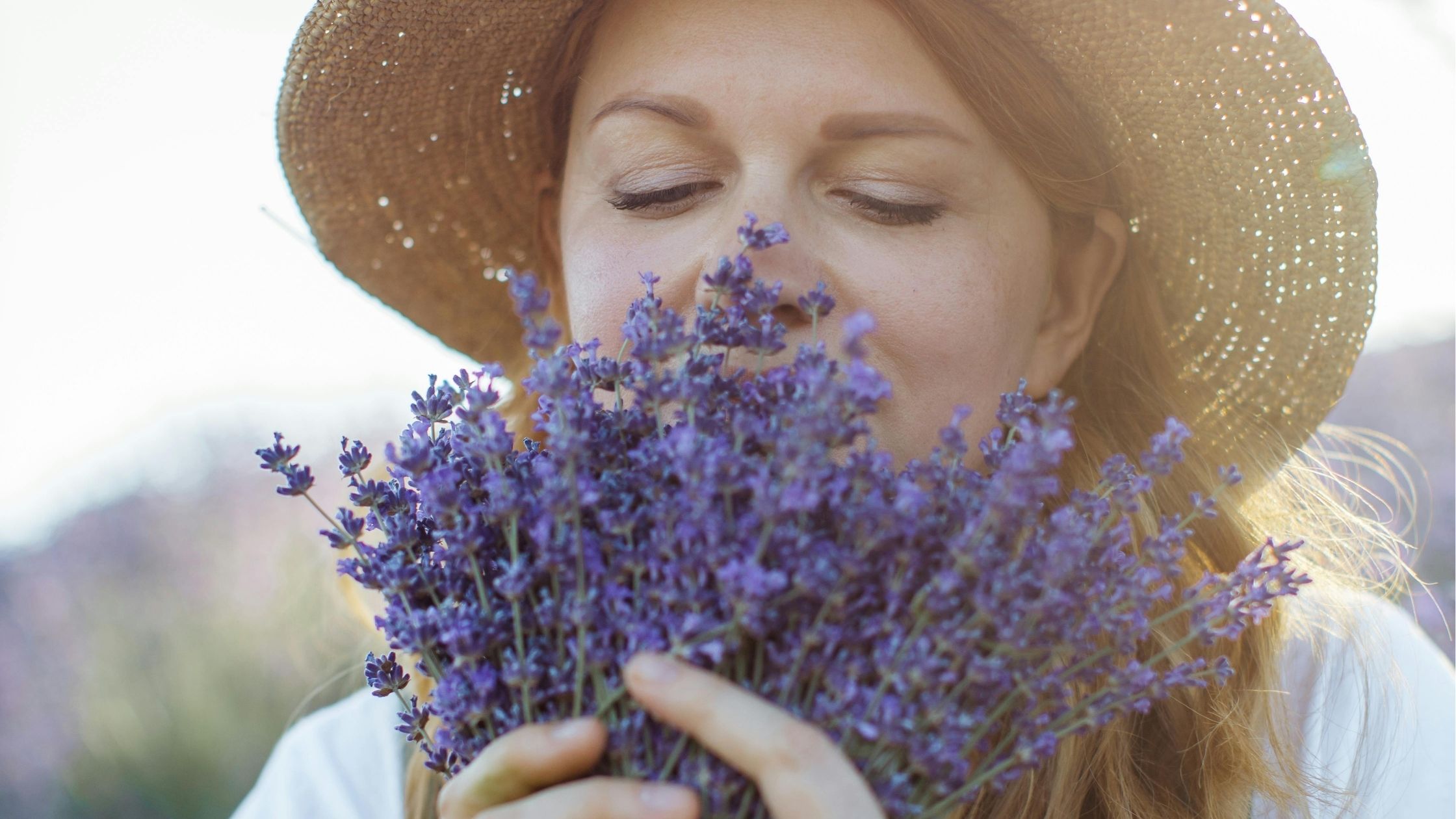 Serene woman in wide-brimmed straw hat inhaling the fragrance of fresh lavender flowers with eyes closed in peaceful enjoyment outdoors