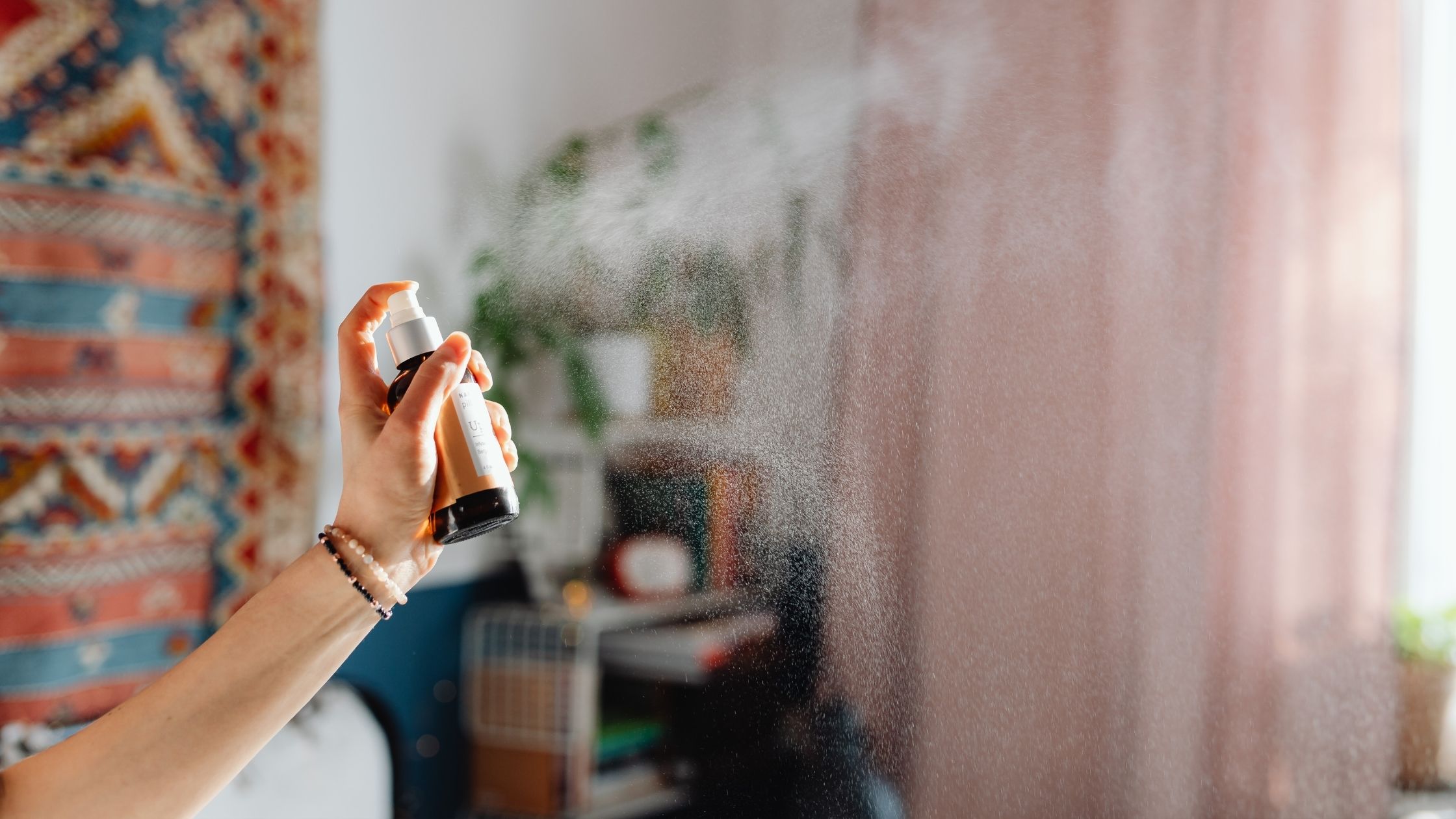 Hand holding and spraying a fine mist from an amber aromatherapy room spray bottle in a bright, serene indoor setting
